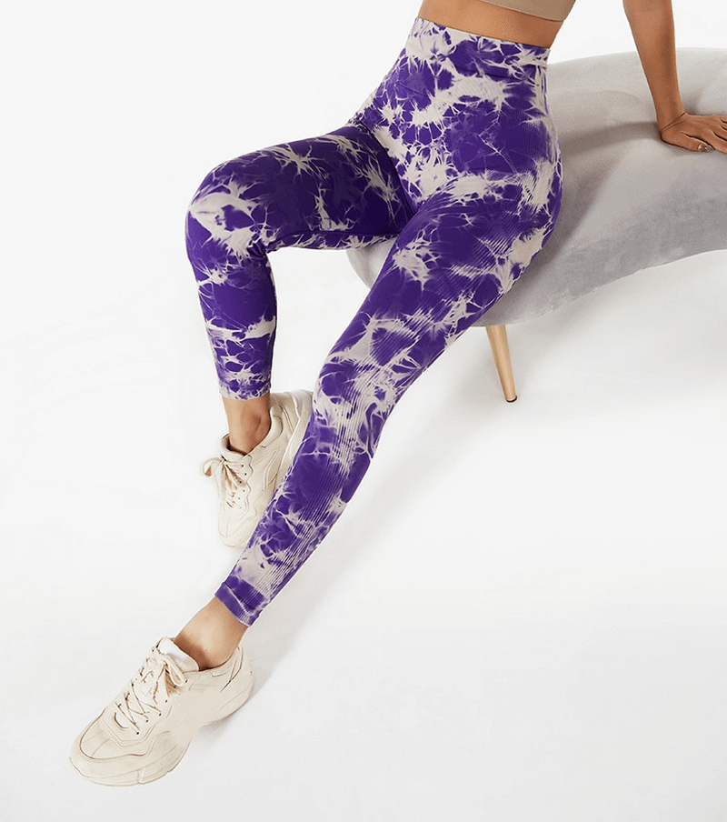Purple tie-dye seamless elastic women's high waist fitness leggings worn by model sitting on a chair, paired with white sneakers